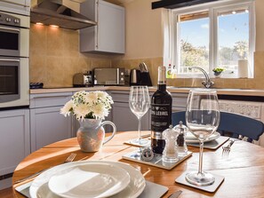 A kitchen with a dining table set for two, featuring a wine bottle, salt and pepper shakers, and a vase with flowers. - The Cottage - UK35177 (Buckland Brewer, near Bideford)