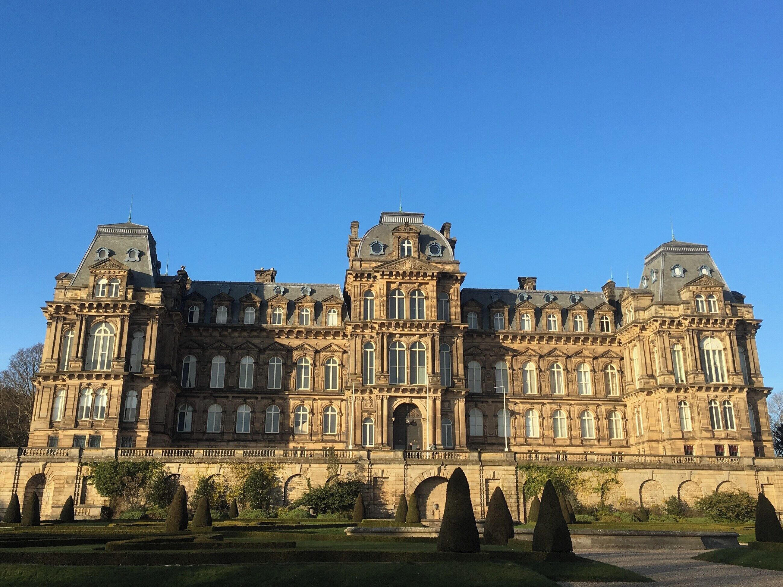 Un gran edificio histórico de múltiples alas y fachada simétrica, con detalles arquitectónicos ornamentales y un jardín formal en primer plano.