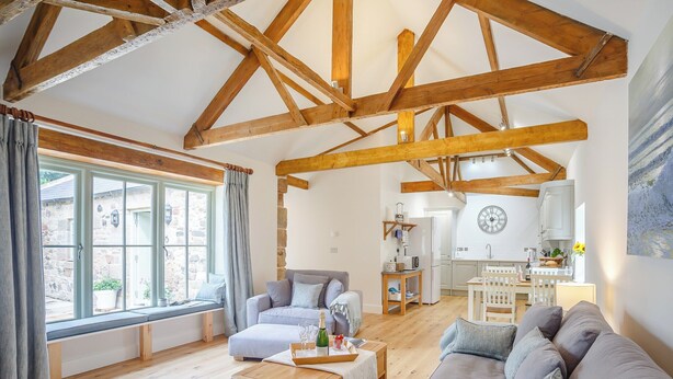 Un salon spacieux avec un plafond haut, des poutres apparentes, un grand canapé gris, une table basse en bois et une vue sur la cuisine et la salle à manger.