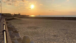 Una playa con un muelle que se adentra en el agua, un atardecer en el fondo y un mar tranquilo.