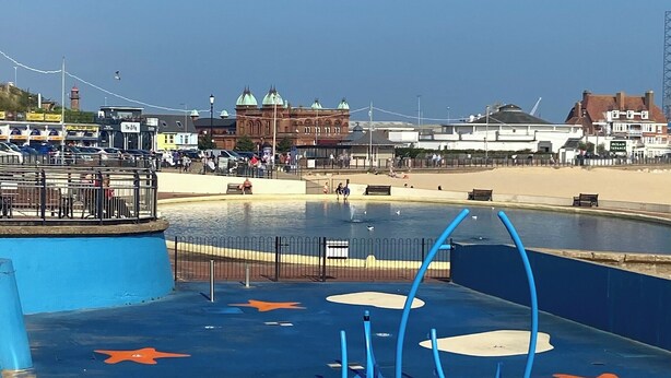 Zona frente al mar con una estructura de juego azul, una playa de arena y un edificio histórico al fondo.