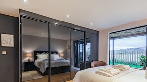 A bedroom with a large mirror, a bed, a nightstand, a chair, and a view of a green landscape. - The Barn at Whitefields (Charlton)