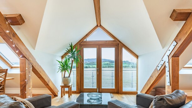 A loft-style room with a sloped ceiling, a wooden staircase, a seating area with leather sofas, a coffee table, and a potted plant. - The Barn at Whitefields (Charlton)