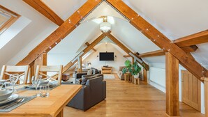 A spacious attic room with wooden beams, a dining table set for two, a sofa, and a television. - The Barn at Whitefields (Charlton)