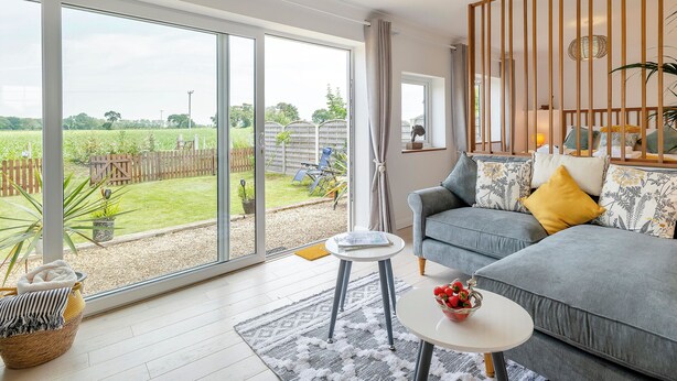 A living room with a sofa, a coffee table, and a view of a garden through large windows. - 1 bedroom accommodation in Ingham, near Stalham (Ingham, near Stalham)