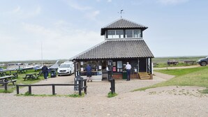 En lille toetages bygning med et tag af fliser og vejrhane, omgivet af picnicborde og en grussti.