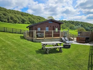 Eine Holzjagdhaushälfte mit einer Terrasse, einem Whirlpool und einem Picknicktisch in einer grünen Wiese, im Hintergrund ein bewaldeter Hügel.