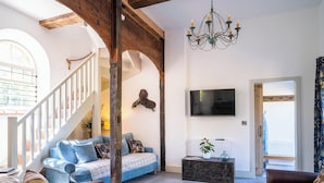 Un salon spacieux avec un plafond haut, un escalier en bois, un téléviseur fixé au mur et une grande fenêtre.