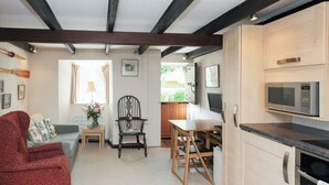 A compact living space with a kitchenette, a wooden dining table, a red armchair, a small side table, and a window with curtains. - 1 bedroom accommodation in Batson (Batson)