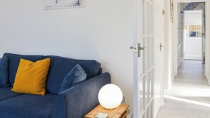 Un soggiorno moderno con un divano blu, un cuscino giallo e una lampada bianca rotonda su un tavolino in legno. Si intravede un quadro al muro.