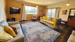 A living room with a brick wall, a flat-screen TV, a yellow sofa, a beige sofa, and a patterned rug. - Bethanga - Short walk to the heart of Robe -Pets Welcome - Outside Only (Robe)