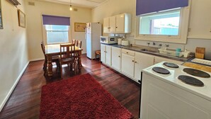 A compact kitchen with a dining area, wooden chairs, a red rug, a white stove, and a window with blinds. - Bethanga - Short walk to the heart of Robe -Pets Welcome - Outside Only (Robe)