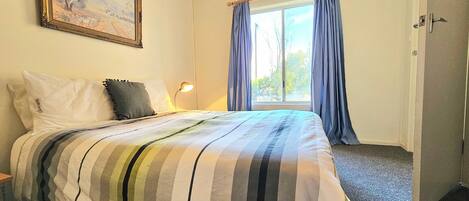 A hotel room with a bed, a nightstand, a lamp, a window with curtains, and a painting on the wall.