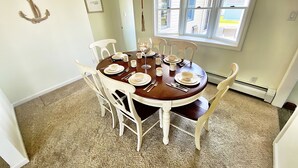 A dining room with a round table set for six, white chairs, a chandelier, and a window with blinds.
