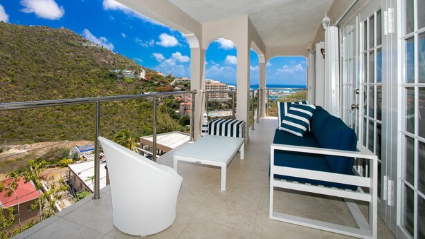 A balcony with a white table, a blue sofa, and a white chair. There is a view of a mountainous landscape and a body of water in the background. - Centrally located minutes away from great beaches, town center and French side. (Philipsburg)