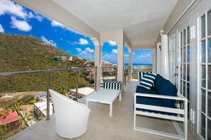 Un balcon avec une table blanche, un canapé bleu et une chaise blanche. On peut admirer une vue sur un paysage montagneux et une étendue d’eau en arrière-plan.