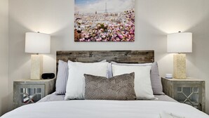 Habitación de hotel con una cama, dos mesitas de noche con lámparas, un cabecero de madera y un cuadro de un paisaje urbano en la pared.