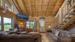 Interior acogedor de una cabaña de troncos con techo de madera, televisor montado en la pared, un cartel que dice “The Best Place to Be is Together” y vistas del exterior a través de amplias ventanas.