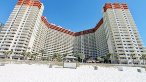 Un hotel de gran altura con varios balcones, una zona de playa con palmeras y un cielo azul despejado.