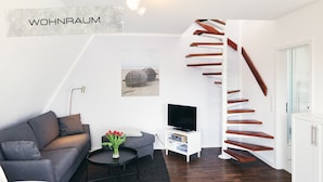 Un salon moderne avec un canapé gris, une table basse noire, un téléviseur posé sur un meuble blanc et un escalier en colimaçon avec des marches en bois.