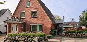 A red brick house with a small porch, a stone wall, and a garden with flowers and shrubs.