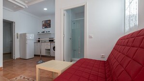 Ein kompakter Wohnbereich mit einem roten Sofa, einem kleinen Holztisch, einer Küchenzeile inklusive Kühlschrank und einer Duschkabine aus Glas.