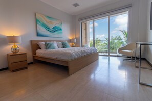 A modern bedroom with a large bed, a desk, and a view of palm trees through the balcony. - EAGLE BEACH-Luxury condo at LeVent with amazing pool and ocean views! (Eagle Beach, Oranjestad)