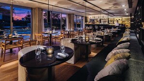 A modern restaurant with tables set for dining, a bar area, and a view of the sunset through large windows. - SLS Buenos Aires Puerto Madero (Buenos Aires)