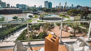 A balcony with two white chairs, a wooden table, and a view of a cityscape including a river and buildings. - SLS Buenos Aires Puerto Madero (Buenos Aires)