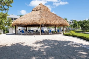 A thatched-roof pavilion with blue chairs and a gravel floor.