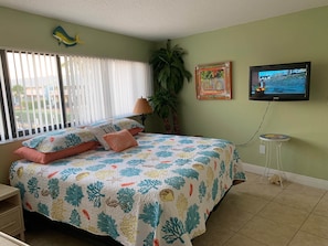 A bedroom with a bed, a TV, a window with blinds, a plant, and a wall with a picture.