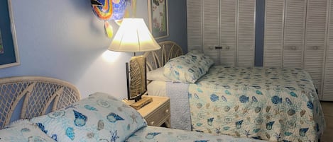 Un dormitorio con dos camas, una mesita de noche, una lámpara, una silla de mimbre y una pared con un cuadro de un pez.