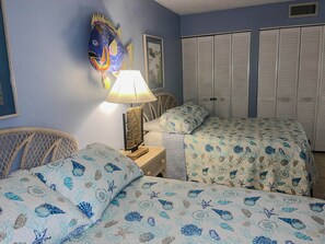Quarto com duas camas, uma mesa de cabeceira, abajur, cadeira de vime e um quadro de peixe na parede.