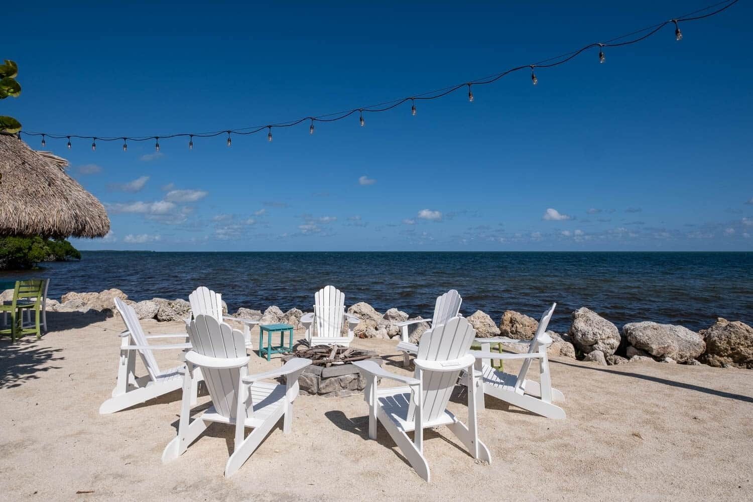 A beach setting with white Adirondack chairs arranged around a small fire pit, overlooking the ocean.