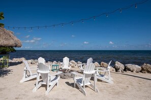 A beach setting with white Adirondack chairs arranged around a small fire pit, overlooking the ocean.