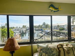 Une chambre avec vue sur un port de plaisance, un poisson décoratif accroché au mur, une lampe en osier et un lit agrémenté d’oreillers à motifs.