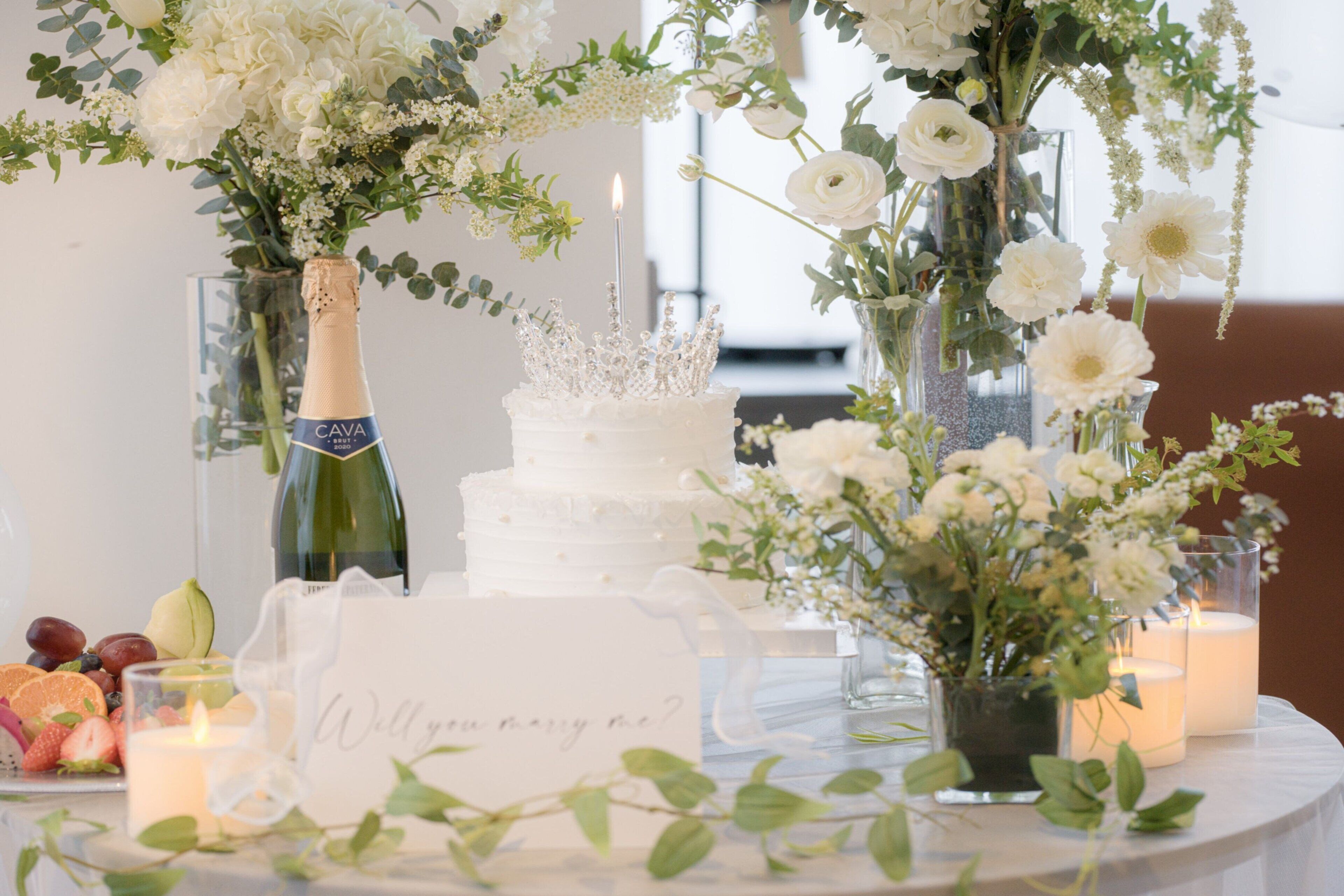 Une table avec un gâteau blanc, une bouteille de cava et une carte sur laquelle est écrit : « Veux-tu m’épouser ? »
