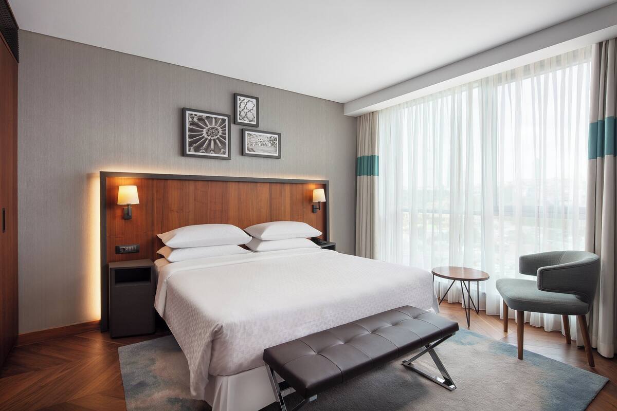 Una habitación de hotel con una cama grande, mesitas de noche, un banco, una silla y vistas a la ciudad a través de cortinas traslúcidas.