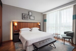 Une chambre d’hôtel avec un grand lit, des tables de chevet, un banc, une chaise et une vue sur la ville à travers de fines rideaux.