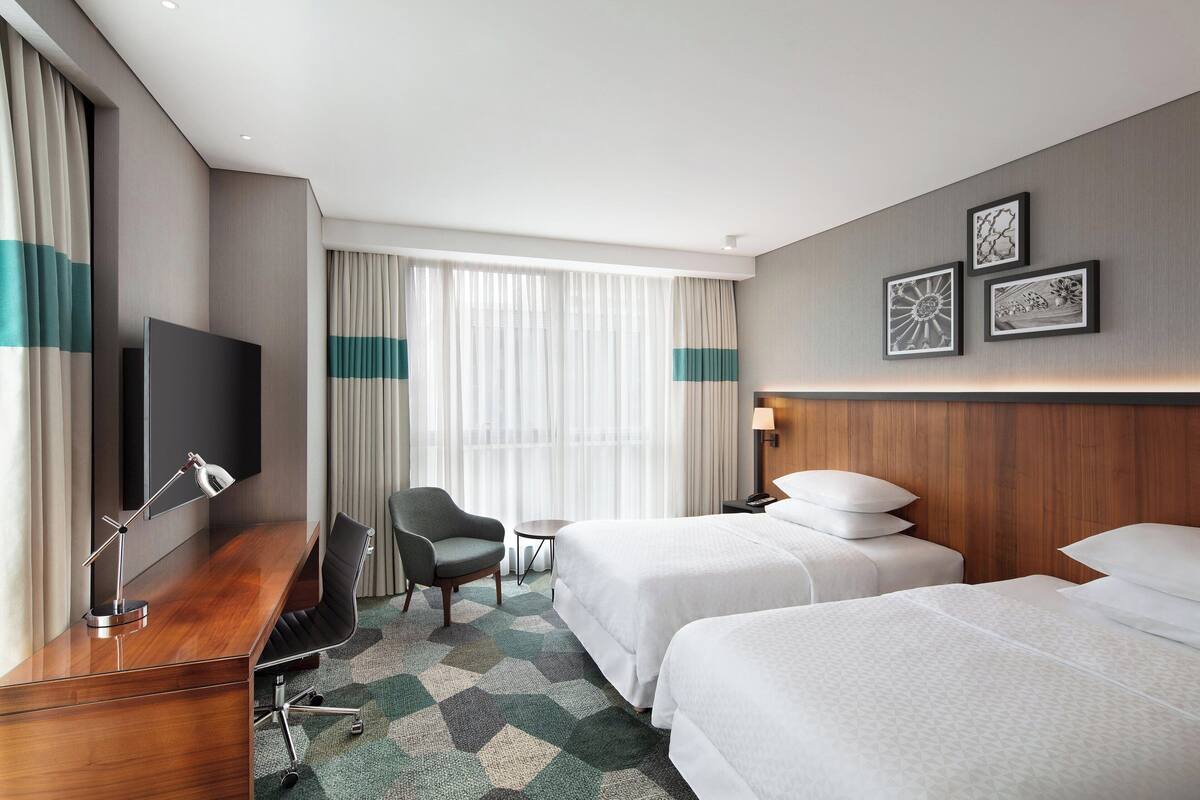 Habitación de hotel con dos camas, un escritorio con silla, televisor y ventana con cortinas.