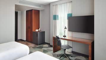 A hotel room with two beds, a desk with a chair, a TV, a wardrobe, and a small kitchenette.