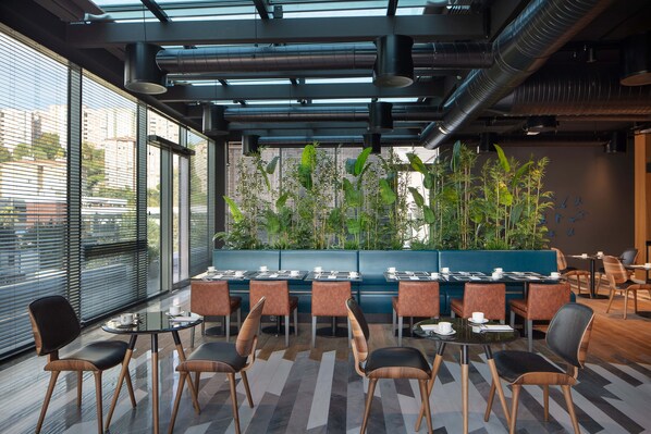 A modern indoor seating area with tables and chairs, large windows, and a green plant wall.