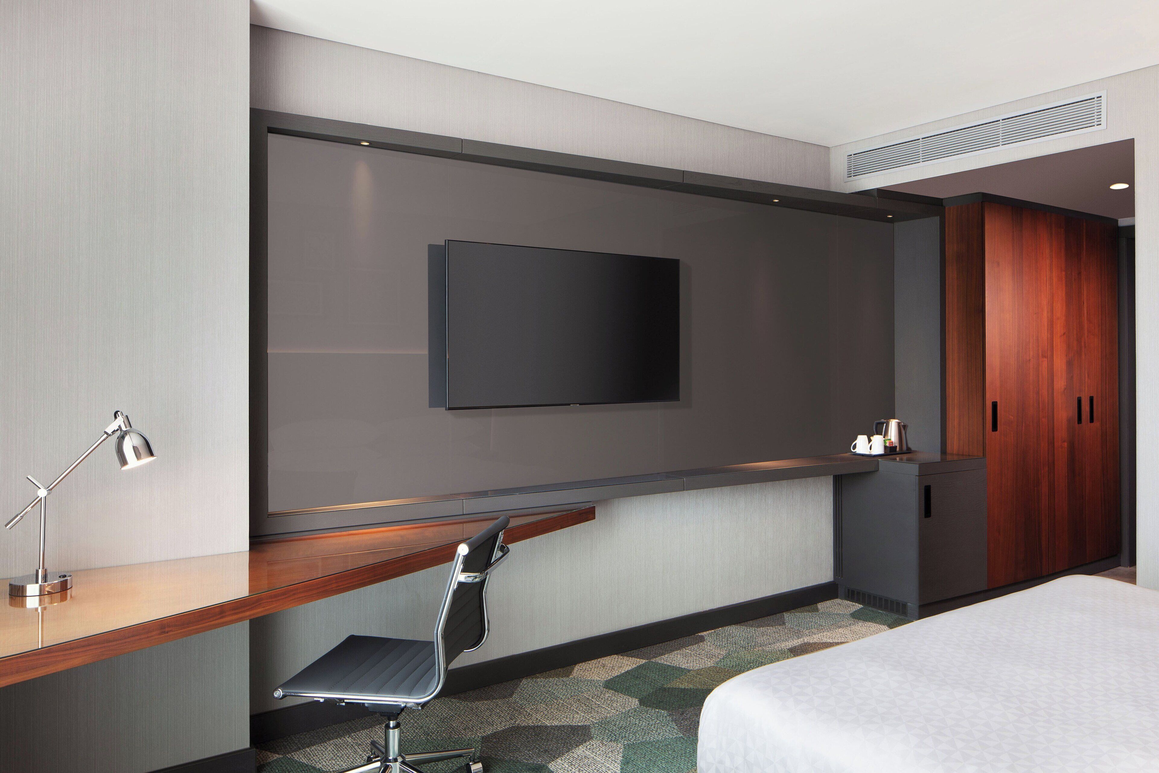 Una habitación de hotel moderna con un televisor de pantalla plana montado en la pared, un escritorio de madera con una silla, una cama y un armario.