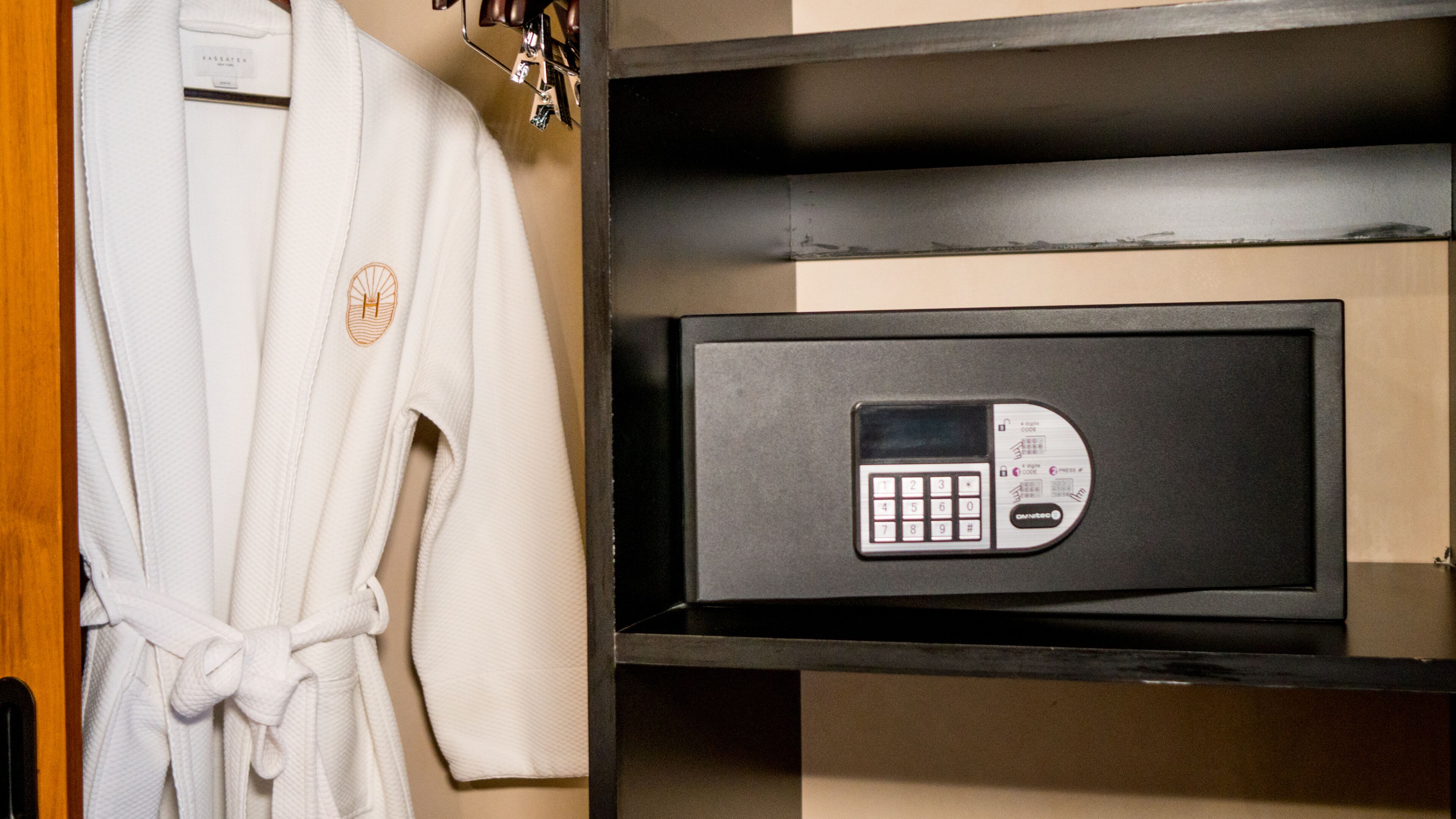 A white robe hanging on a wooden door, a black safe with a keypad, and a shelf with a small item.