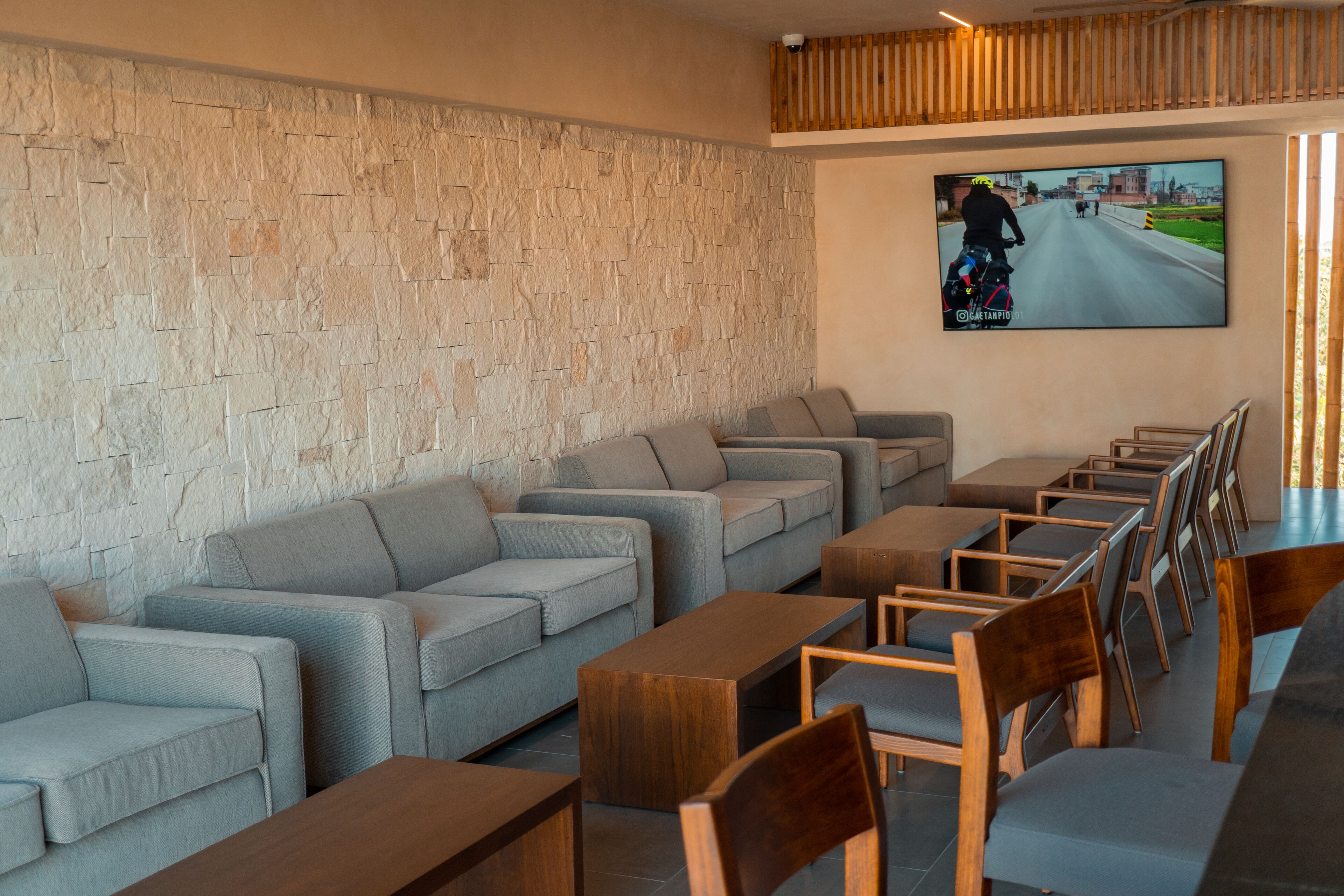 A modern lounge area with sofas and wooden tables, a stone wall, and a mounted TV displaying a cyclist.