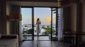 Una sala moderna con un sofá, una mesa de centro y una mesa de comedor. Hay un ventanal con vistas a edificios y al atardecer.