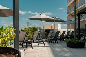 Une terrasse sur le toit, dotée de chaises longues et de parasols, qui offre une vue sur un bâtiment moderne et un ciel dégagé.