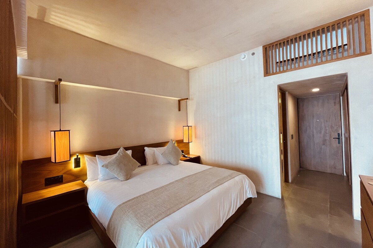 A hotel room with a large bed, bedside lamps, a wooden headboard, a wooden bench, and a door leading to a hallway.