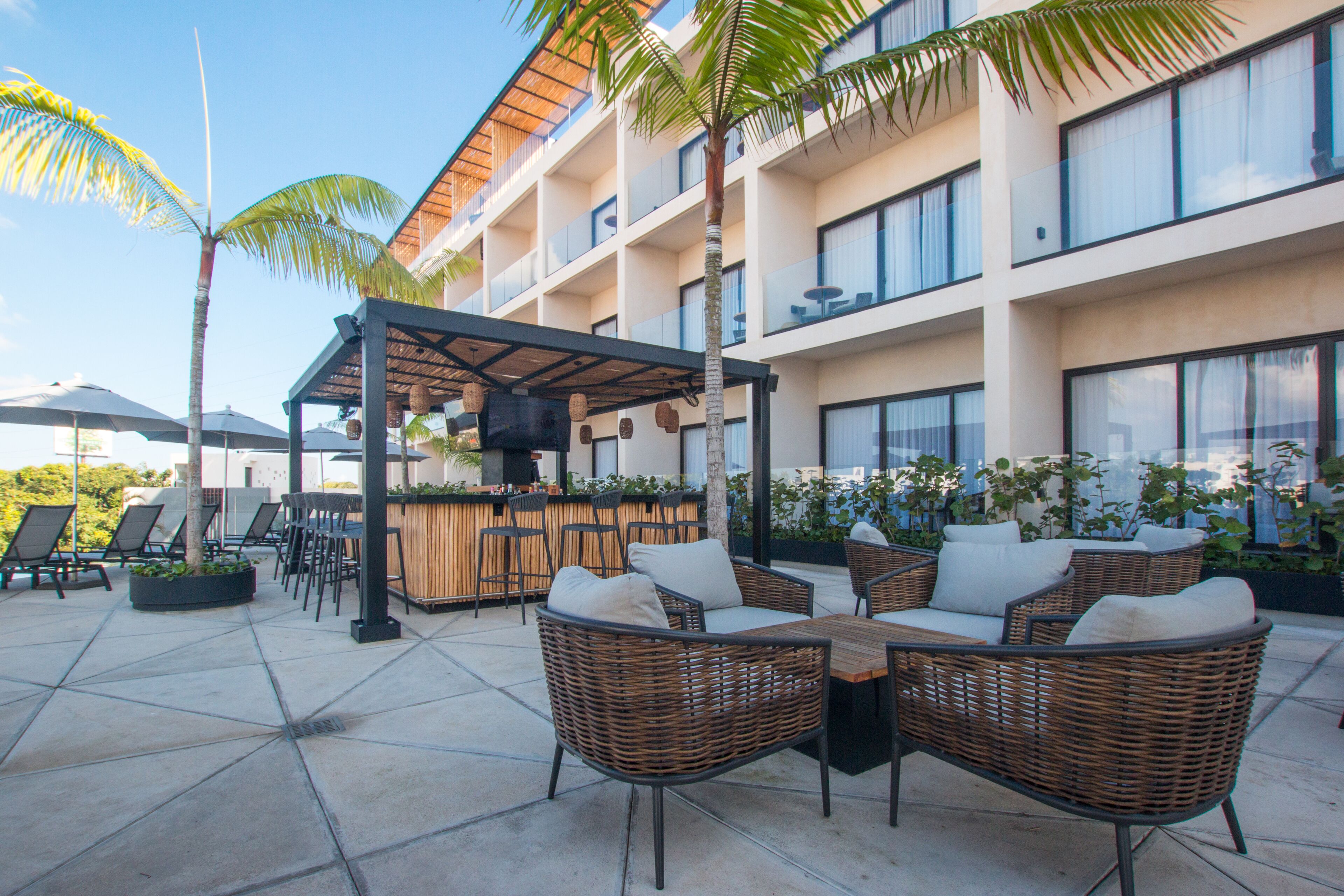 A rooftop bar with wicker furniture and a bar area, surrounded by palm trees and a building with multiple balconies.
