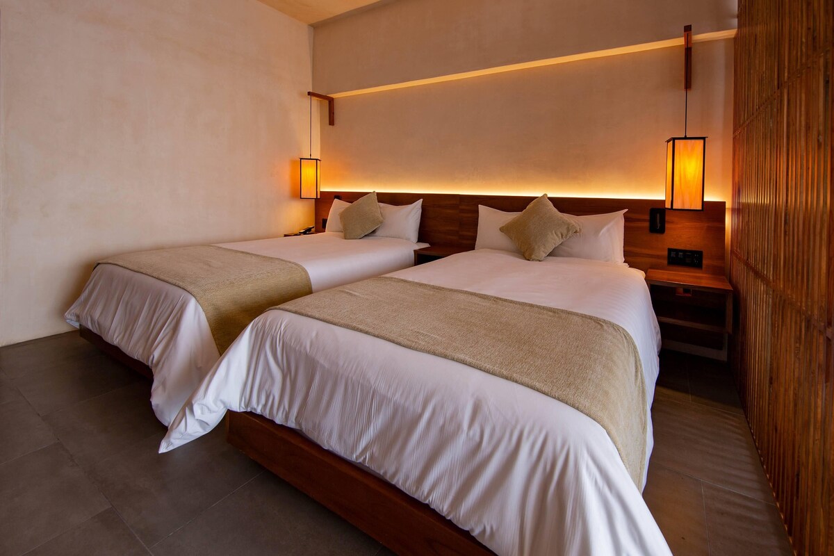 A hotel room with two beds, wooden headboards, bedside tables, and wall-mounted lamps.